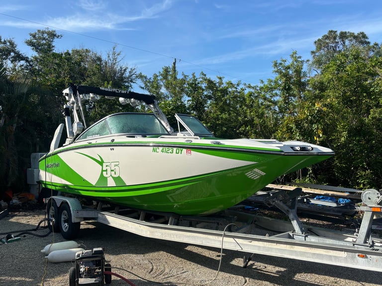 Green and white speedboat with wakeboard tower mounted on trailer in outdoor lot with trees and sky visible